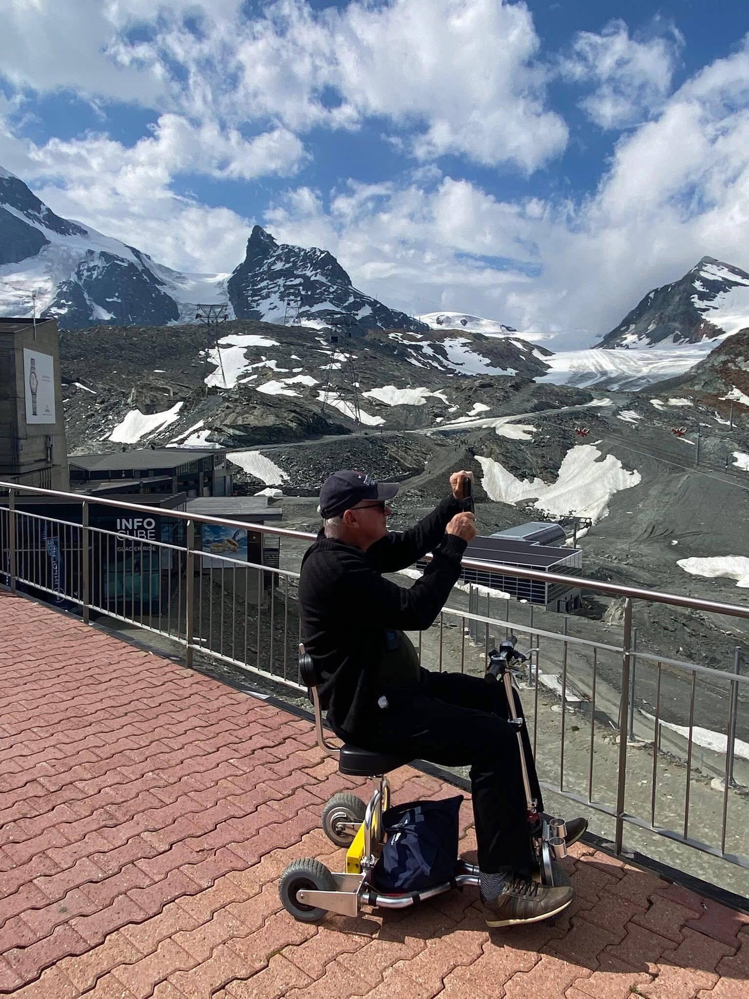Man on mobility scooter oversees snow-capped mountains.